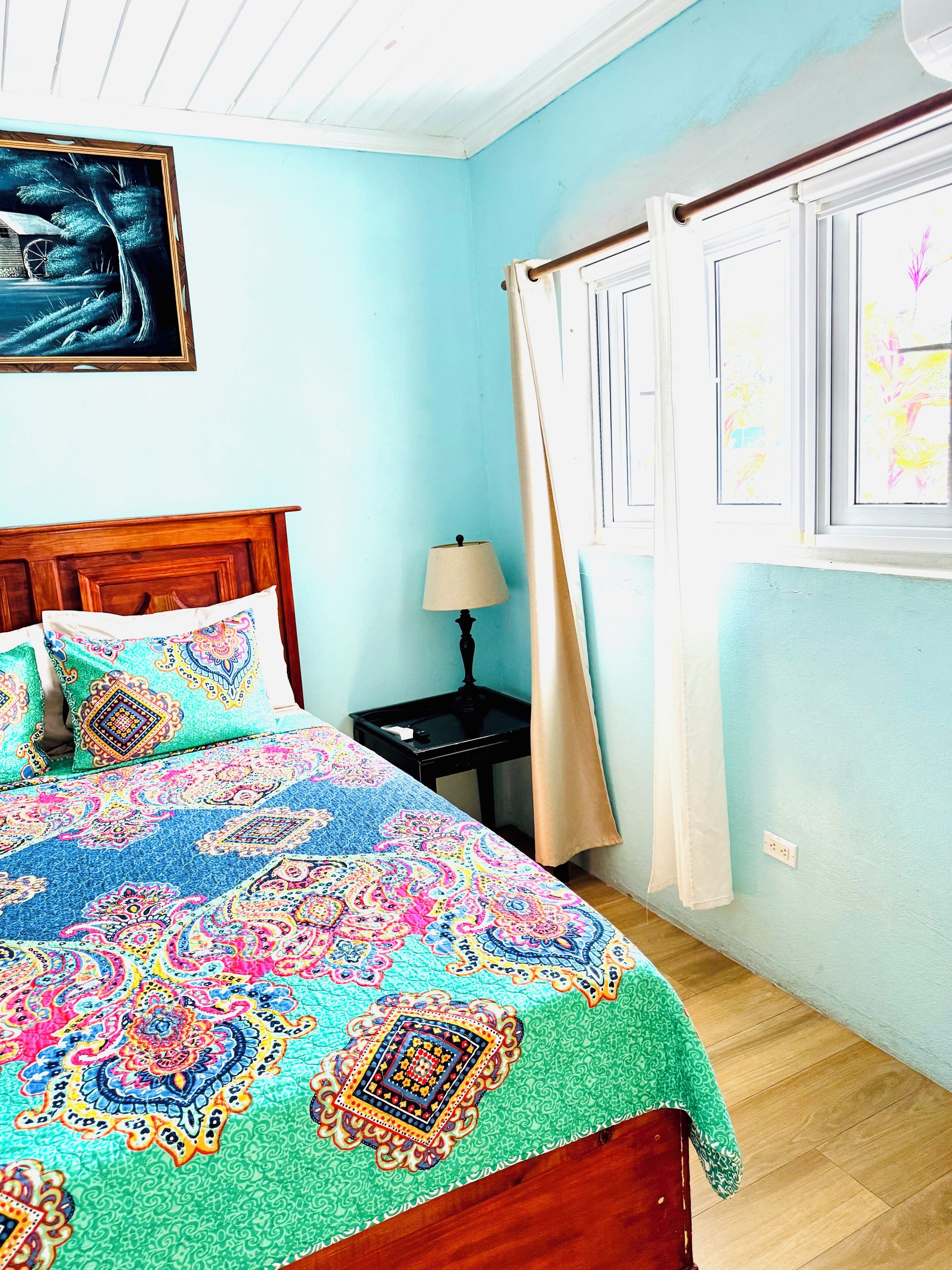 Une chambre à coucher avec un lit, une table de chevet, une lampe, une fenêtre avec des rideaux et un tableau au mur.
