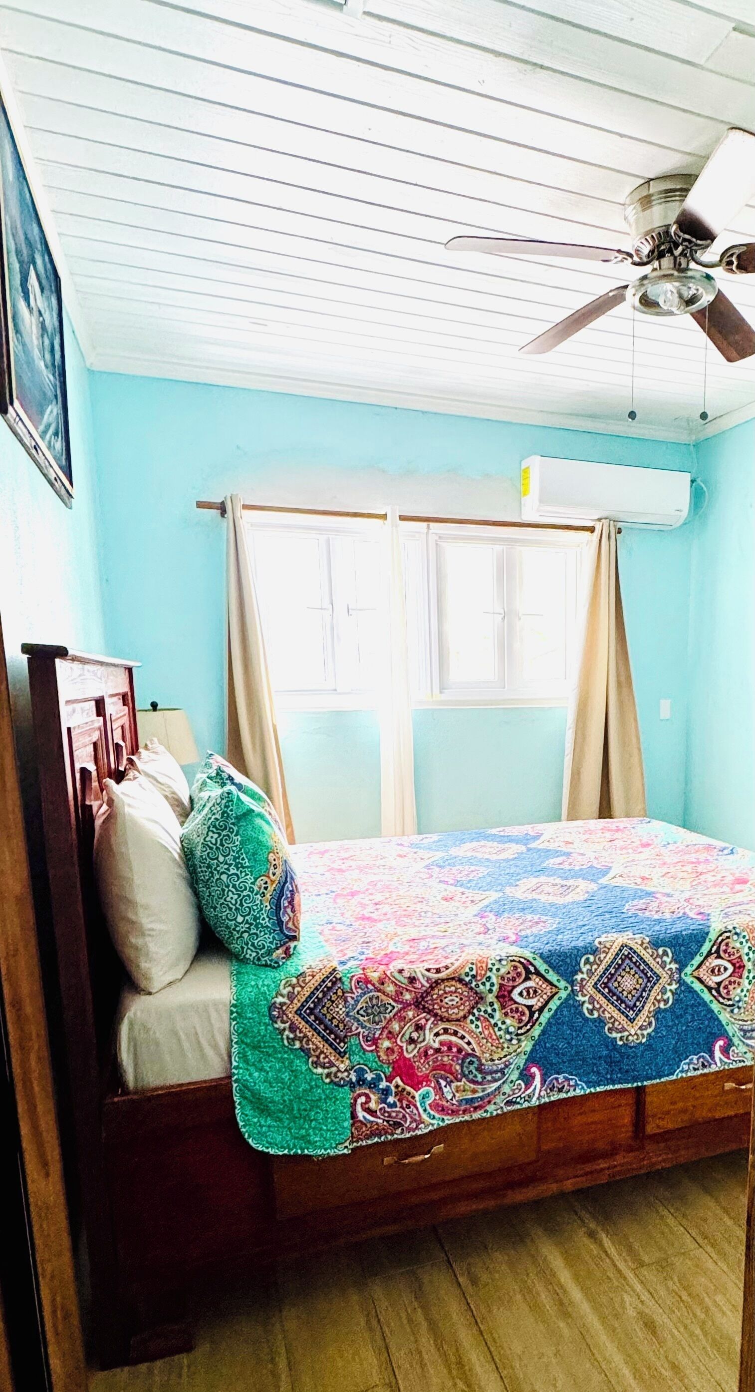 Una habitación con cama, ventilador de techo, ventana con cortinas y aire acondicionado.
