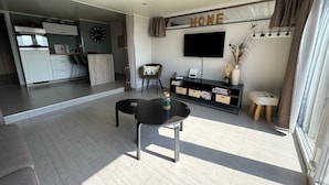 Una sala de estar moderna con un televisor de pantalla plana, una mesa de centro y un sofá. Se ve un área de cocina al fondo.