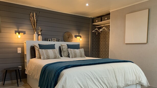 Un dormitorio moderno con una cama grande, dos luces fijadas a la pared, un taburete pequeño y un armario con ropa colgada.