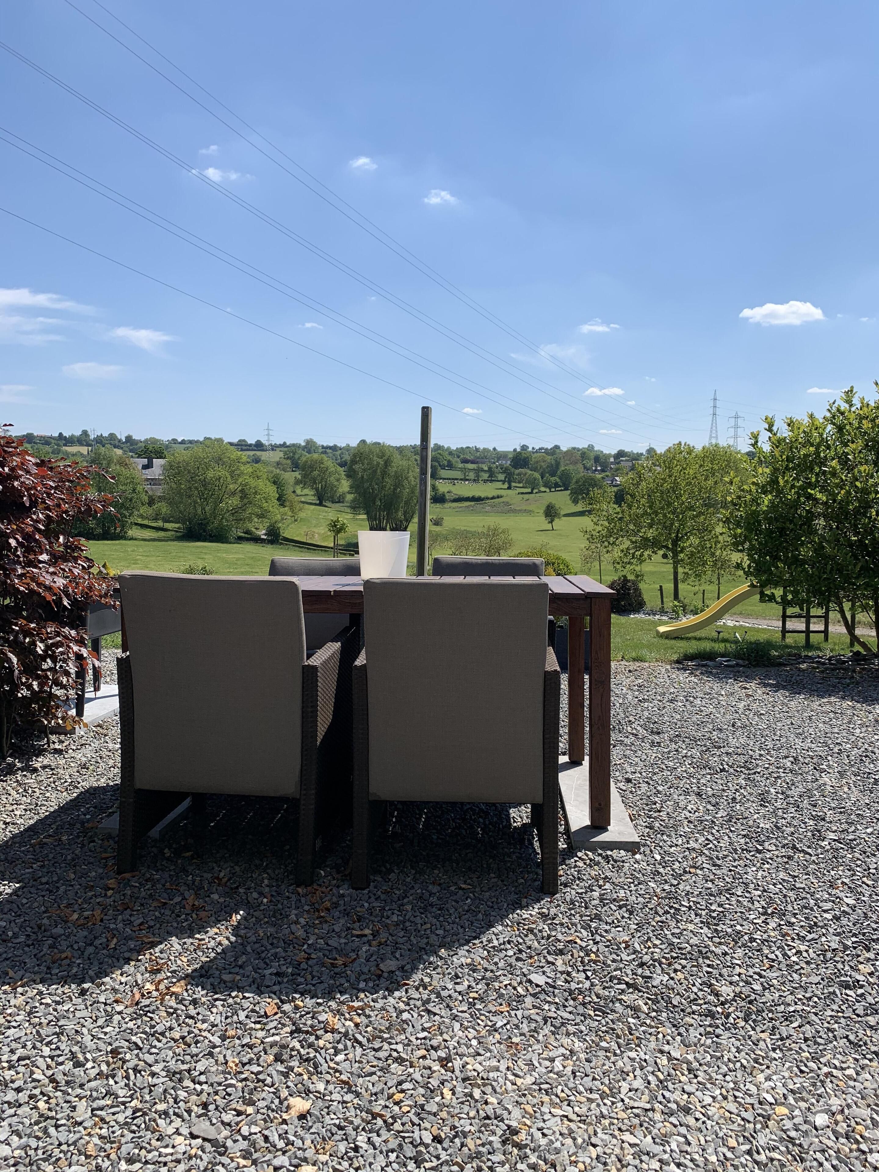 Een terras met een tafel en stoelen, een witte plantenbak, en uitzicht op een groen landschap met bomen en een speeltuin.