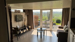 Una sala de estar moderna con un sofá, una mesa de centro y un televisor sobre un mueble bajo. Hay una puerta corrediza de vidrio que da a un balcón con vistas al campo.