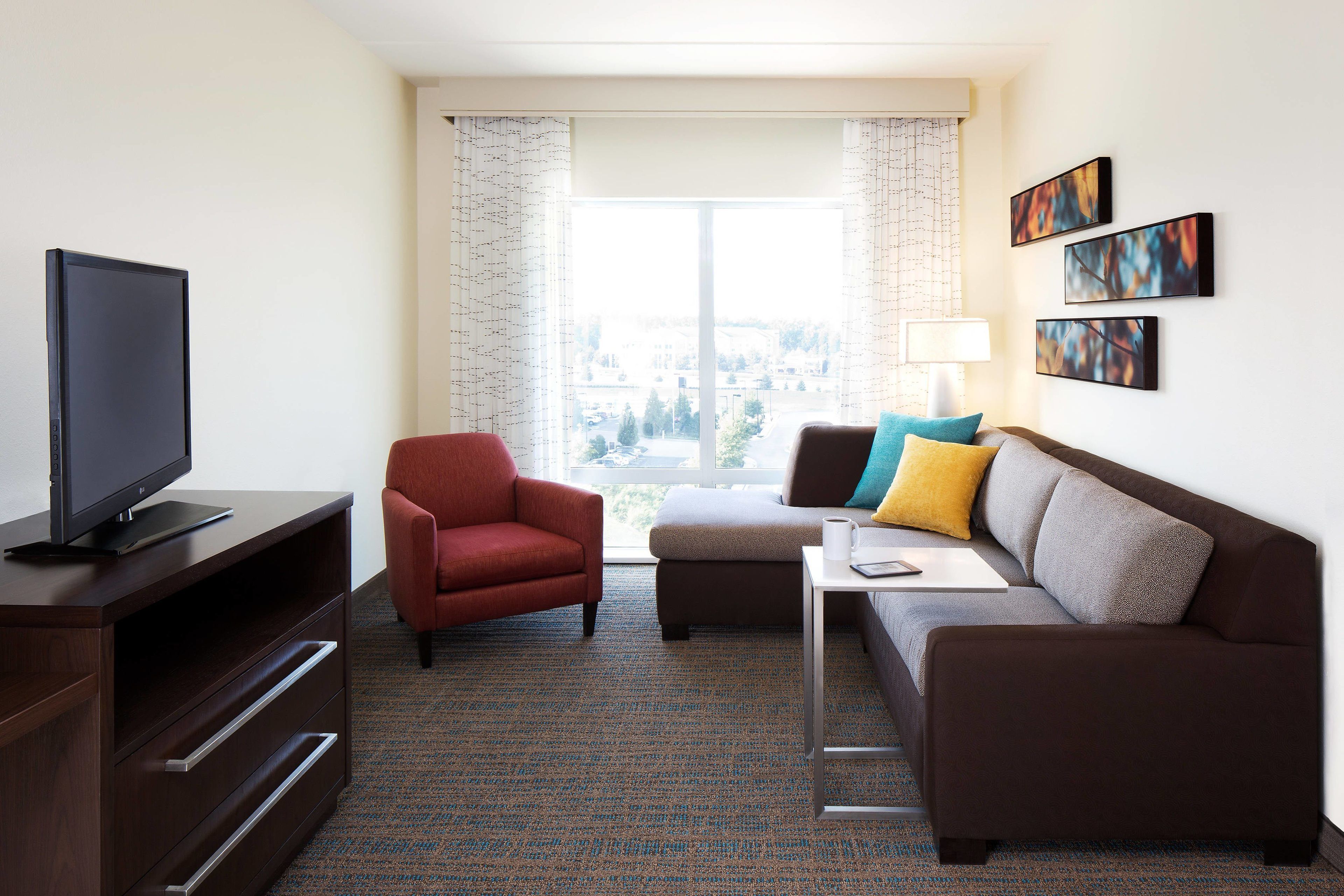 A hotel room with a flat-screen TV, a red armchair, a brown sofa with colorful cushions, a small table, and a window with curtains.
