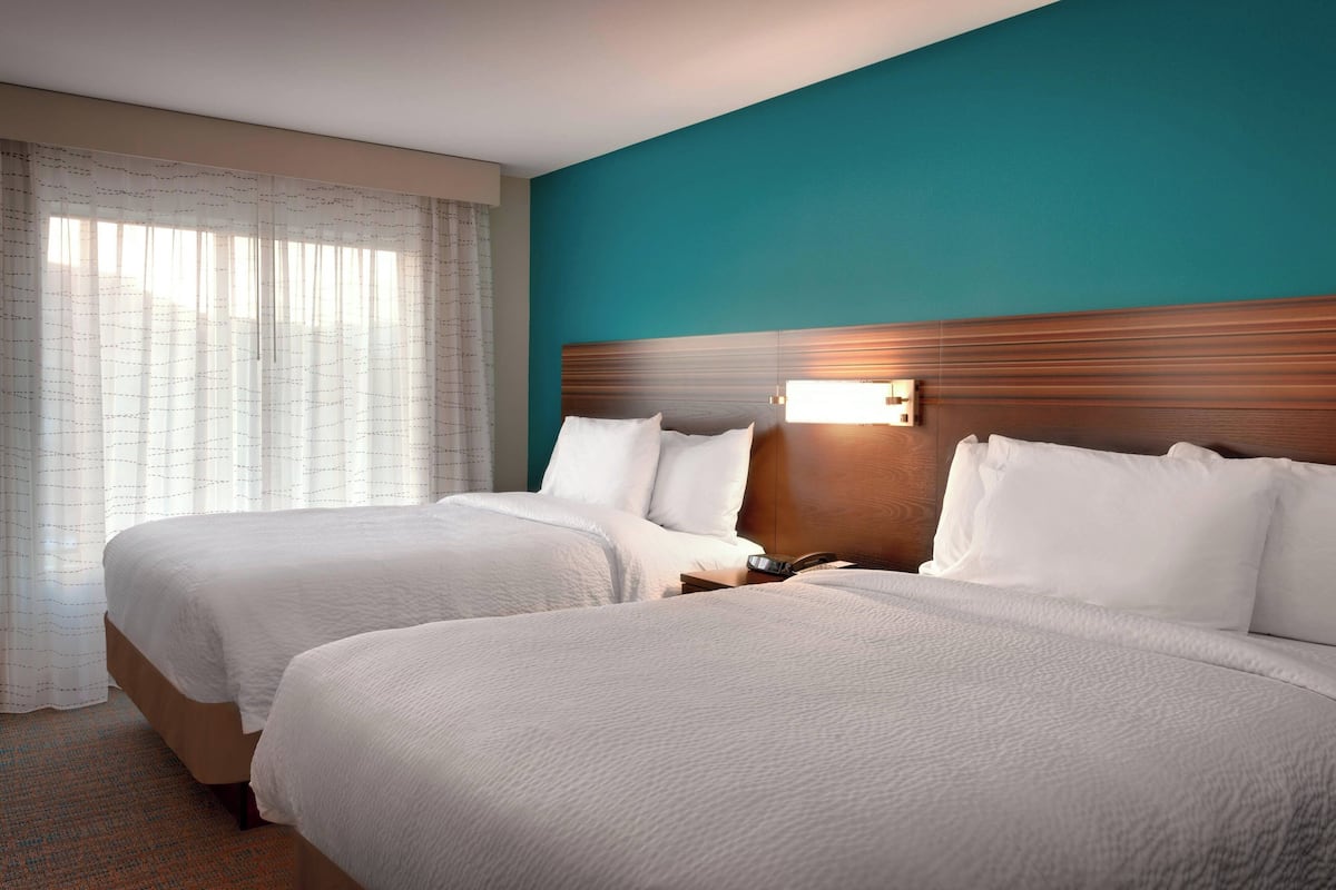Une chambre d’hôtel avec deux lits, une tête de lit en bois et une fenêtre ornée de rideaux légers.