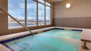 Una piscina interior con agua azul cristalina, un muro de ventanales de vidrio y vistas al exterior de un estacionamiento y montañas.