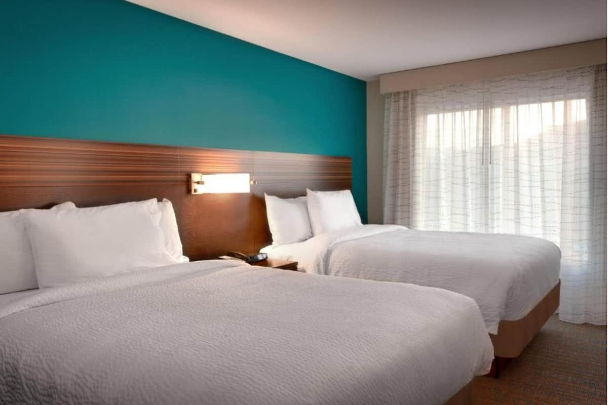 Une chambre d’hôtel avec deux lits, une tête de lit en bois, une table de chevet avec une lampe et une grande fenêtre avec des rideaux légers.