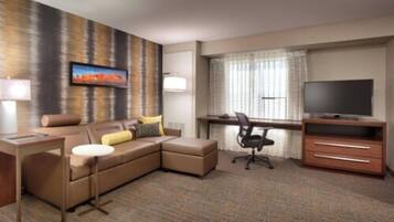 A hotel room with a brown leather sofa, a desk with a computer, a TV, and a large window with curtains.