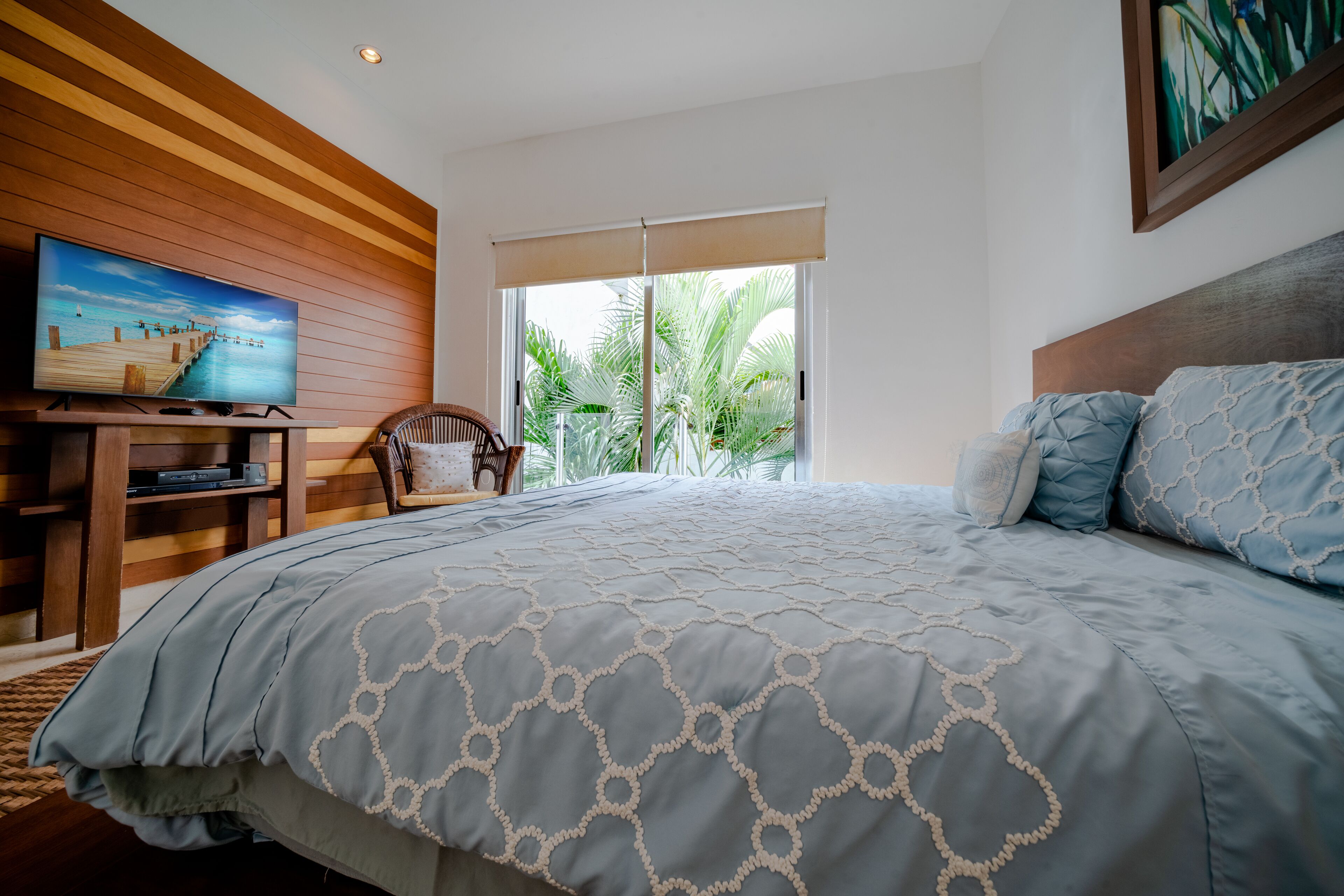 Ein Schlafzimmer mit einem großen Bett, einem Fernseher auf einem Holzgestell, einem Sessel und einem Fenster mit Blick auf Palmen.