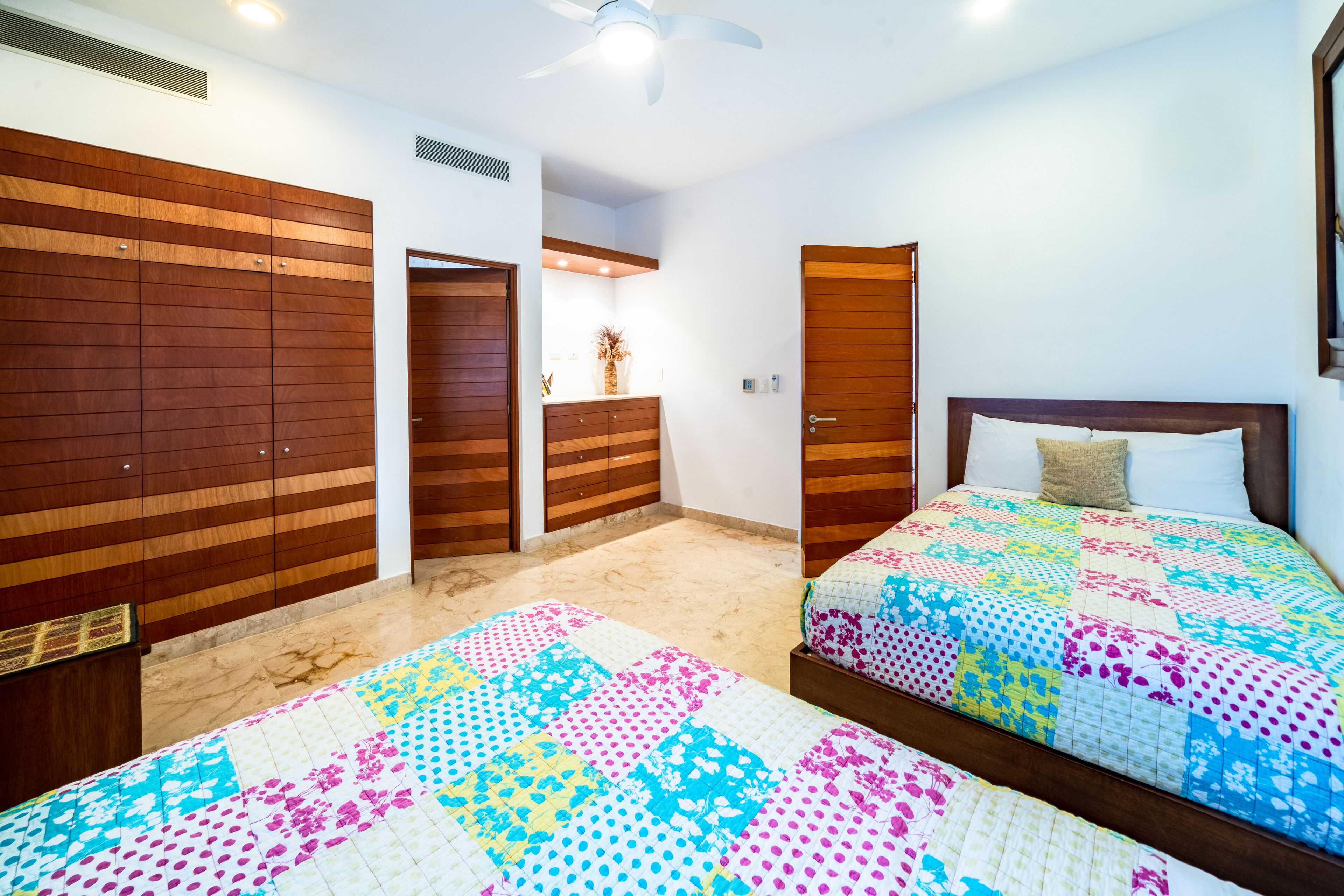 A bedroom with two beds, wooden wardrobes, a ceiling fan, and a bathroom visible through an open door.