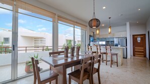 A spacious dining area with a large table and chairs, a kitchen in the background, and large windows offering a view of the surrounding buildings. - Turquesa by NAS (Playa del Carmen)