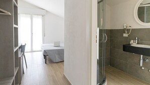 Una habitación de hotel moderna con cabina de ducha de vidrio, una cama con un ordenador portátil, un escritorio con una silla y un ventanal grande.