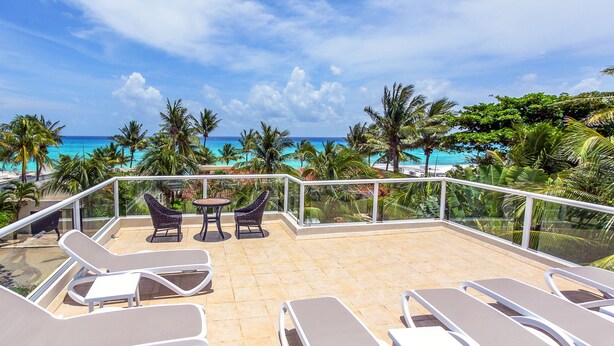 A sunlit outdoor seating area with lounge chairs and a table, overlooking a beach with palm trees and the ocean. - Vista del Mar by NAS (Playa del Carmen)
