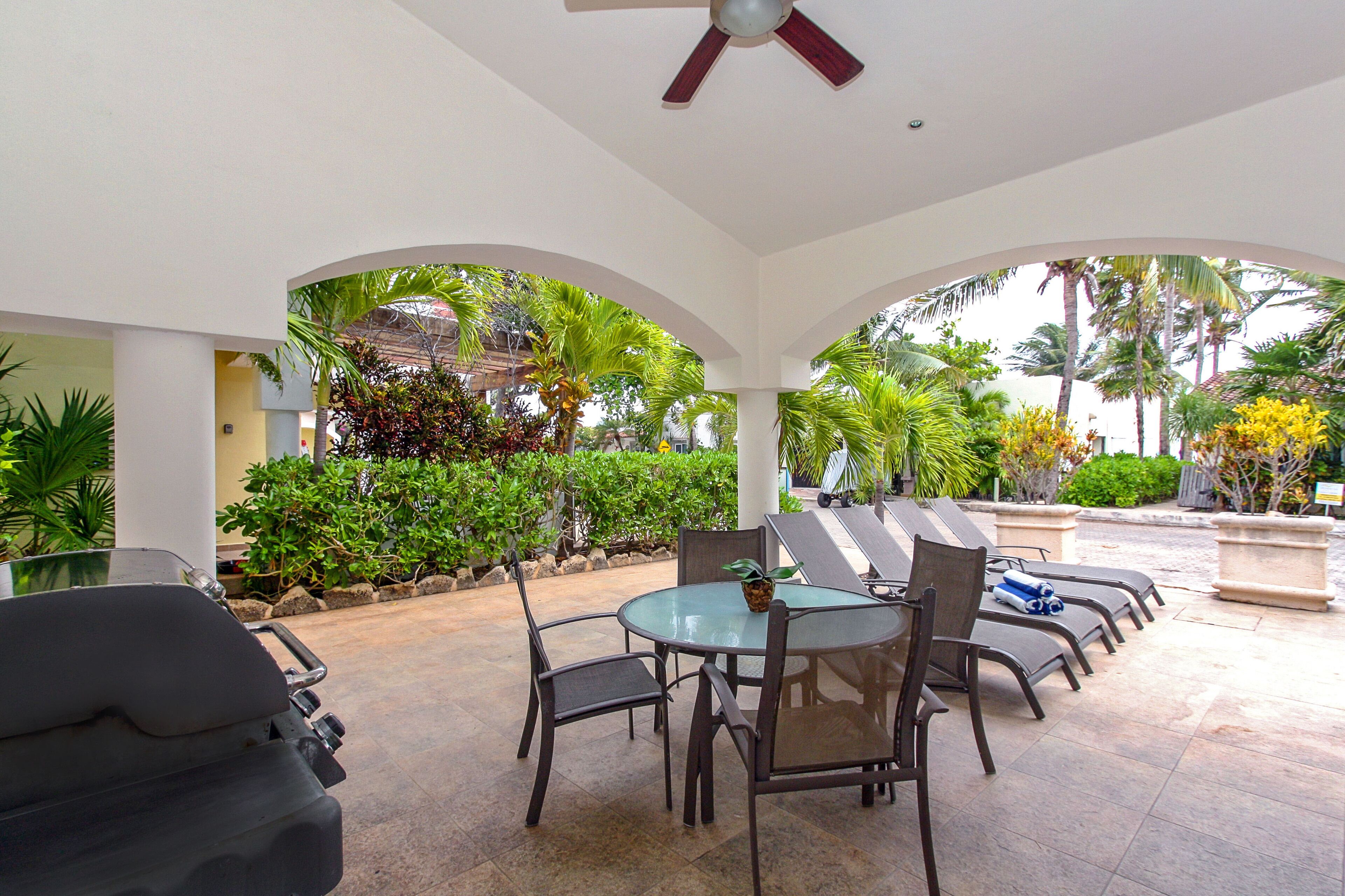 Un patio al aire libre con una mesa redonda y sillas, un ventilador de techo y vistas a plantas tropicales y palmeras.