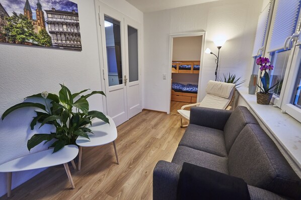 A compact living space with a wooden floor, a grey sofa, a small round table, a potted plant, and a bunk bed in the background.