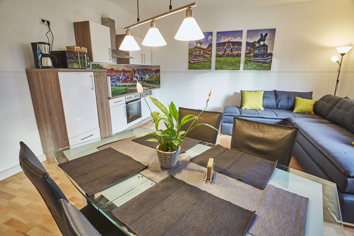 A modern living room with a glass dining table, a sofa, and a kitchen area.