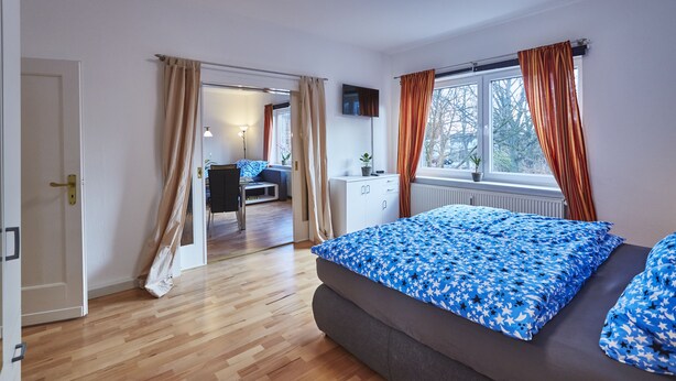 Ein Schlafzimmer mit einem Bett, einem Fenster mit Vorhängen, einem Heizkörper und einer Tür zu einem anderen Raum.