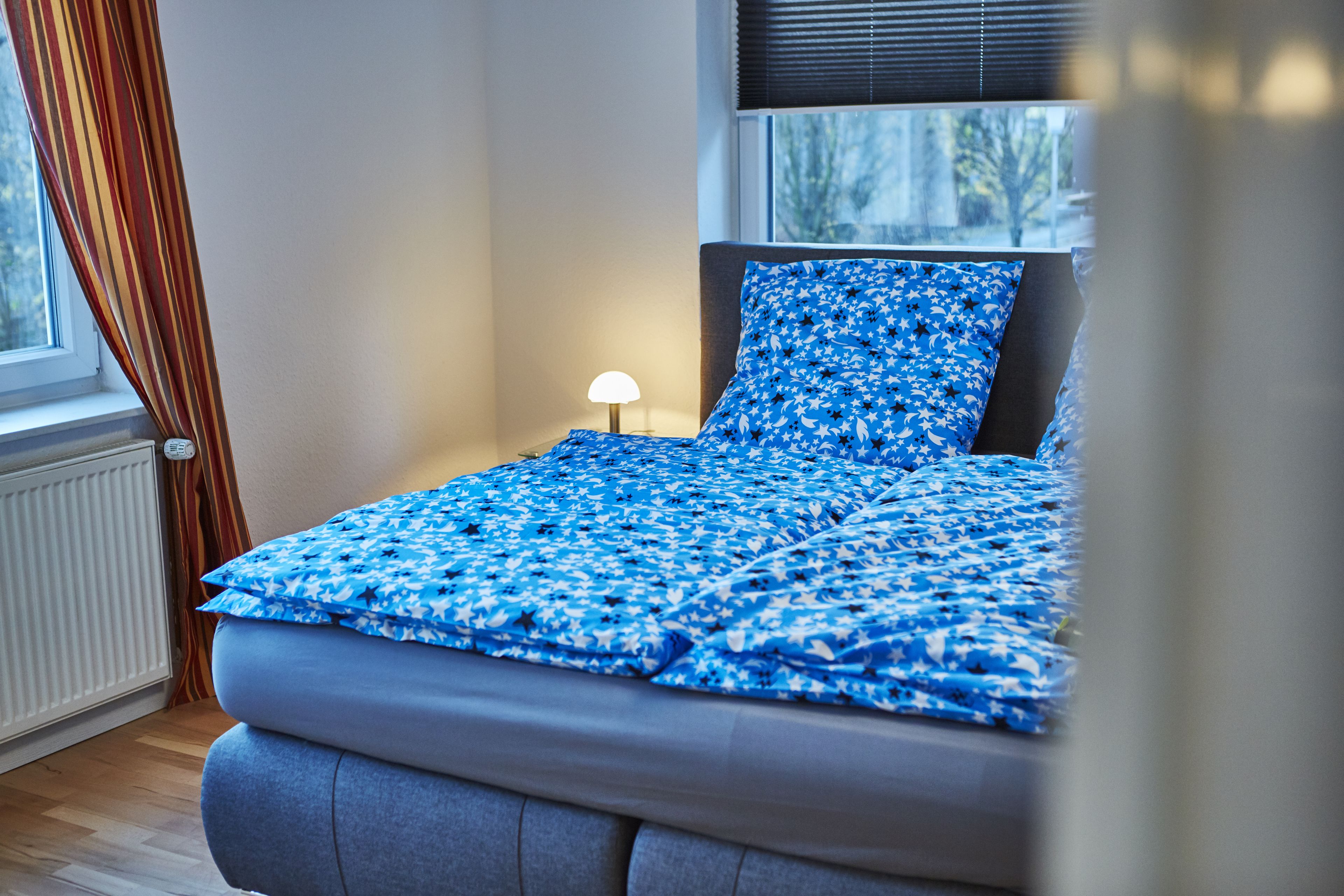 A bed with a blue floral bedspread and a matching pillow, a bedside lamp, and a window with blinds.