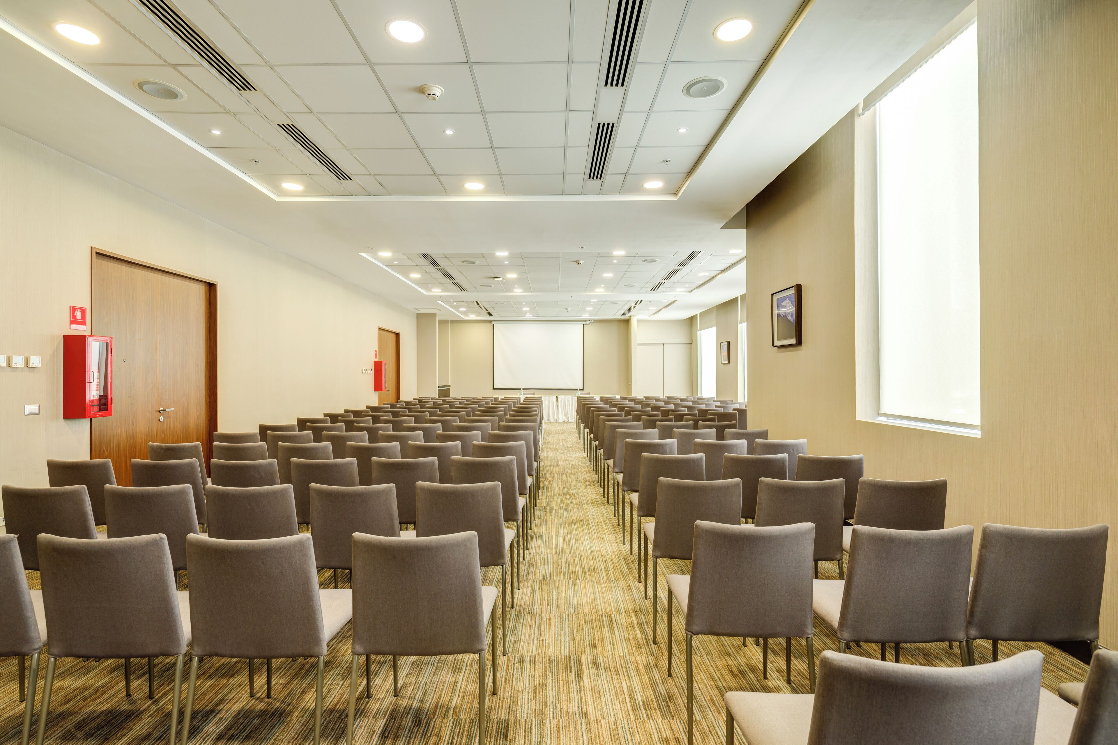 A conference room with rows of chairs, a carpeted floor, a projector screen, and a fire extinguisher.