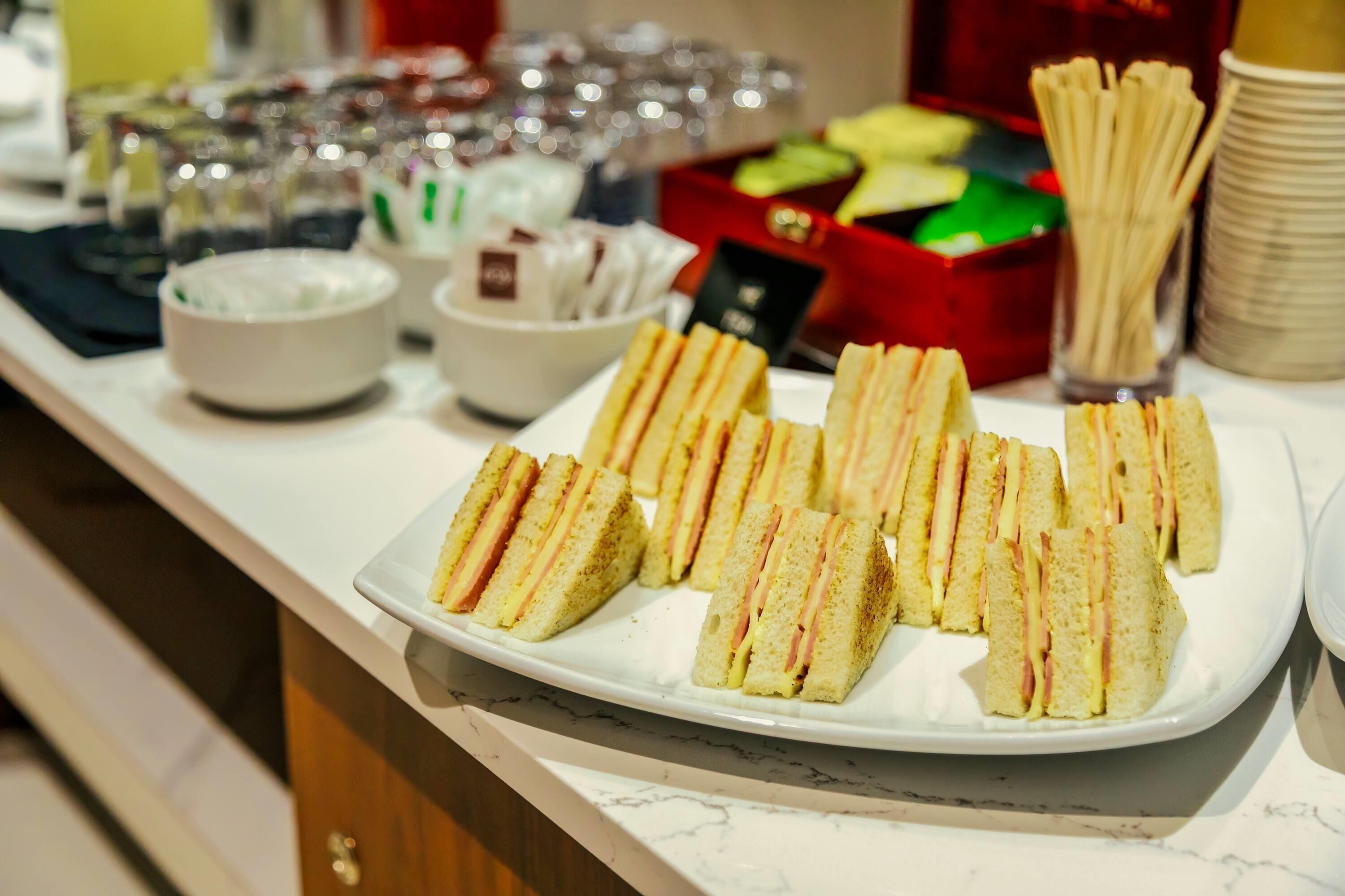 Una mesa de buffet con sándwiches, recipientes con salsas y una caja roja con utensilios.