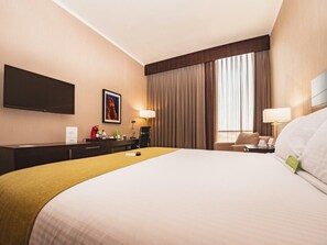 Habitación de hotel con una cama grande, un escritorio con silla, un televisor de pantalla plana montado en la pared y una ventana con cortinas.