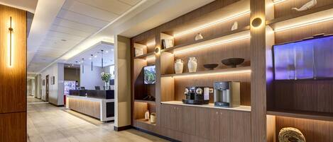 A modern hotel lobby with a reception desk, wooden shelving with decorative items, and a coffee machine.
