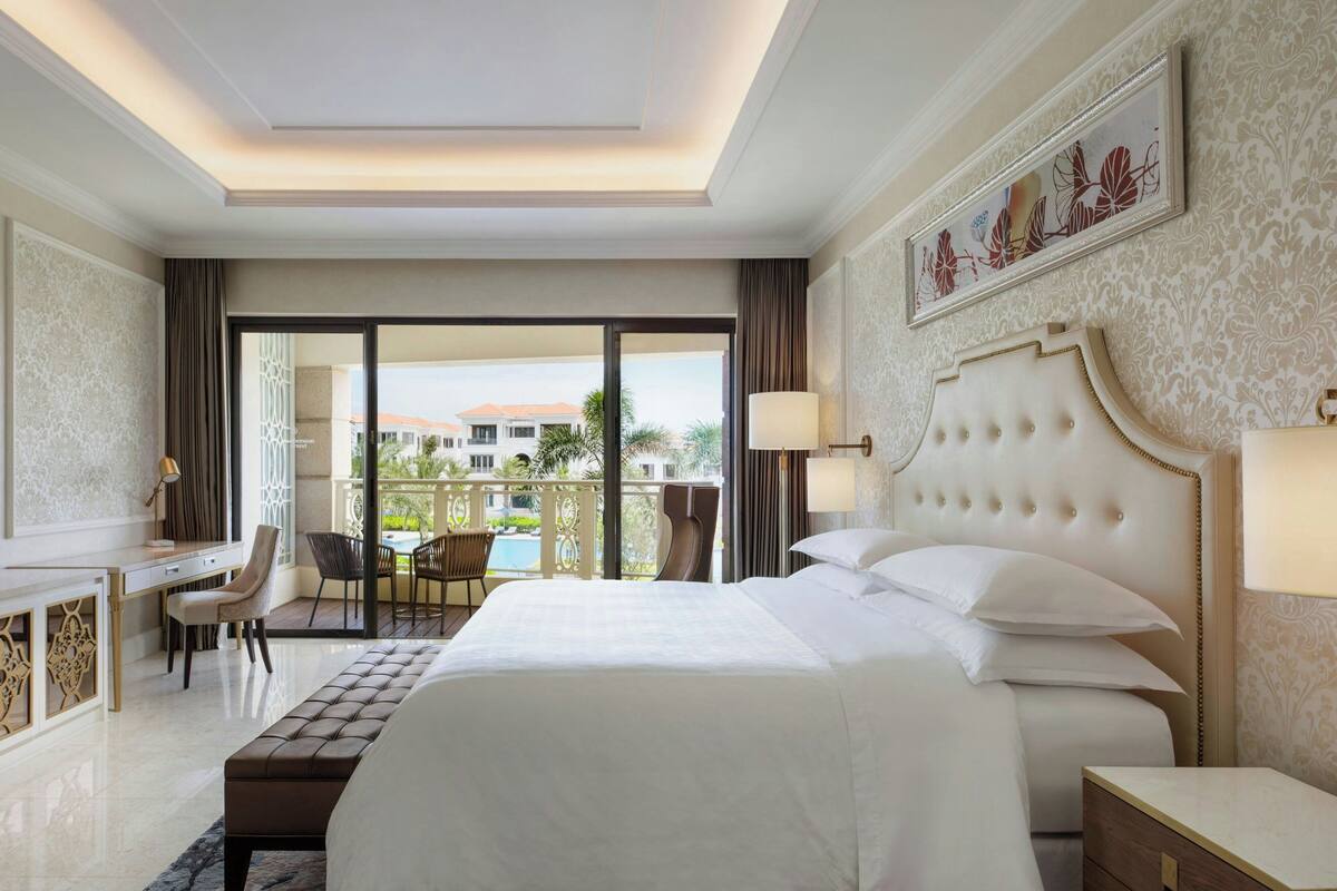 A hotel room with a large bed, a desk, a chair, and a balcony with a view of buildings and palm trees.