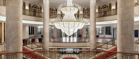 A grand lobby with a large chandelier, marble columns, and a central staircase with red carpet.