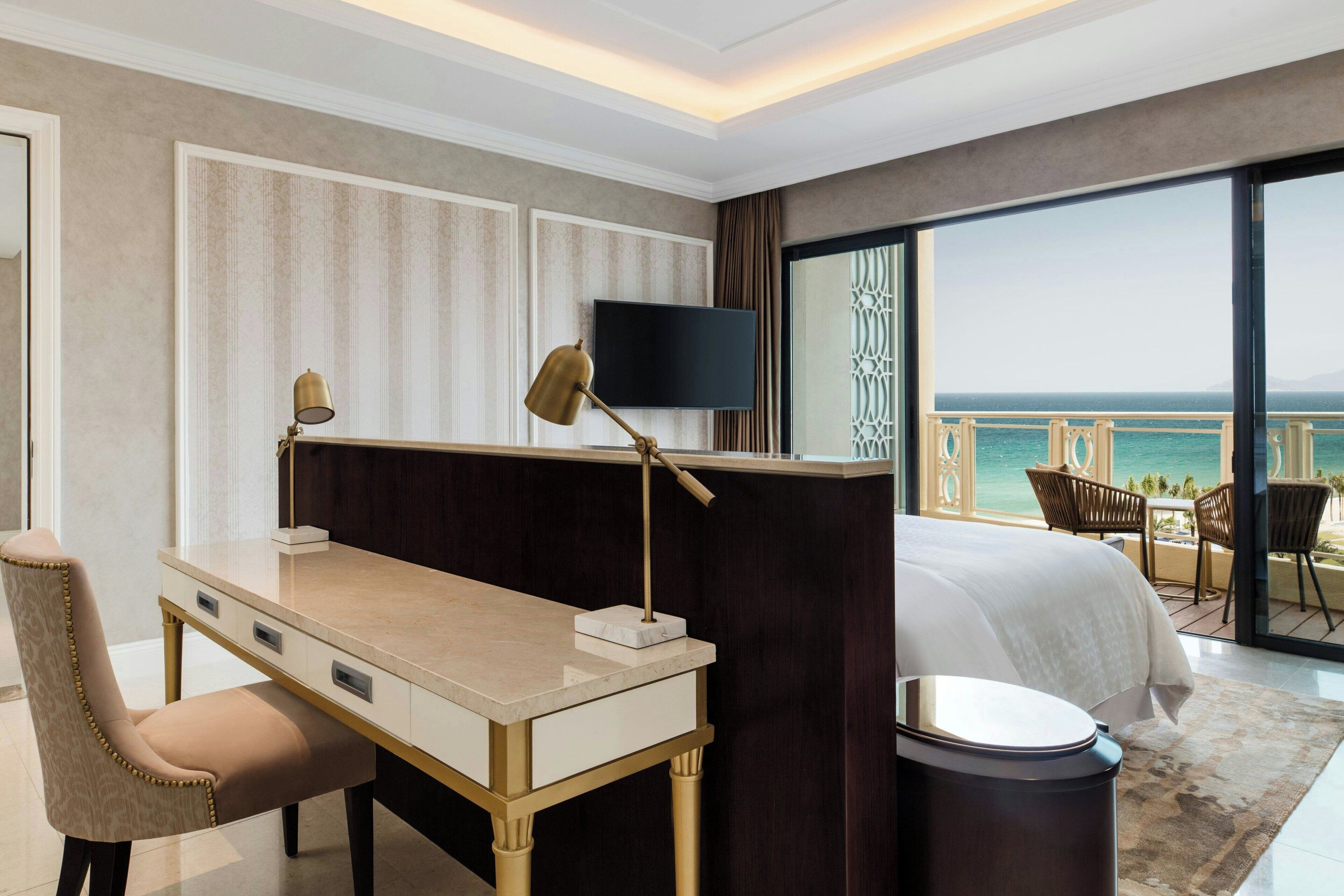 A hotel room with a bed, desk, chair, TV, and a balcony with a view of the sea.
