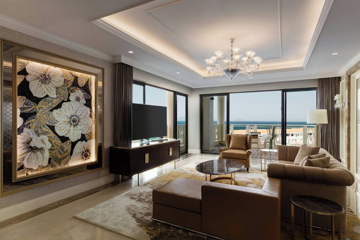A spacious living room with a large flat-screen TV, comfortable seating, a chandelier, and a large window offering a view of the sea.
