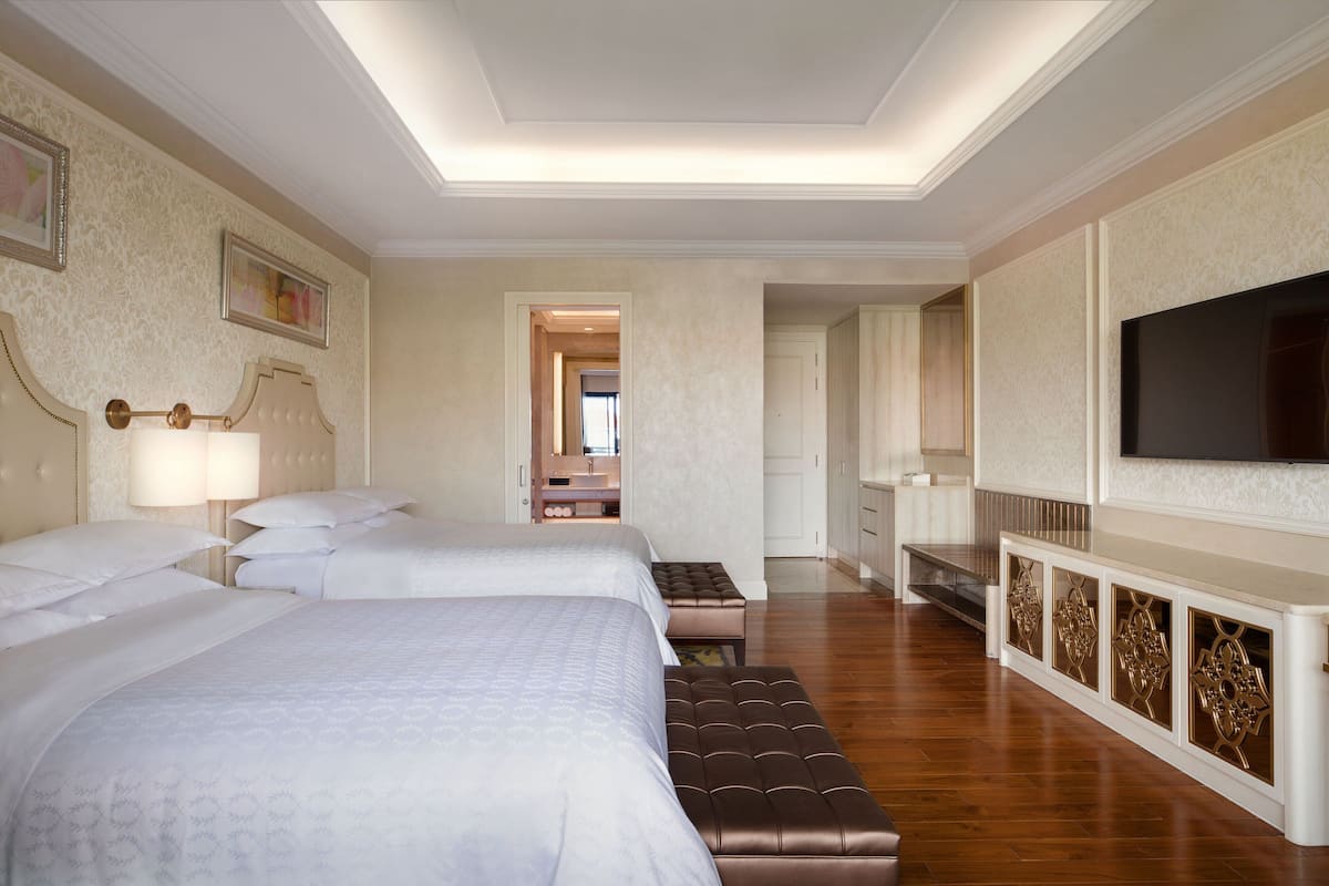 A hotel room with two beds, a TV, a vanity with a mirror, and a hallway leading to another room.