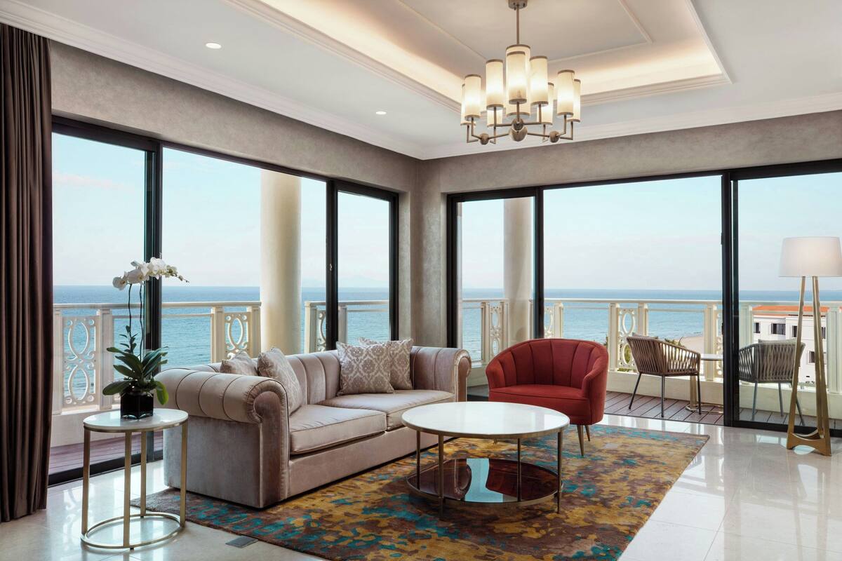 A spacious living room with a large window overlooking the sea, a red armchair, a beige sofa, and a small round table.