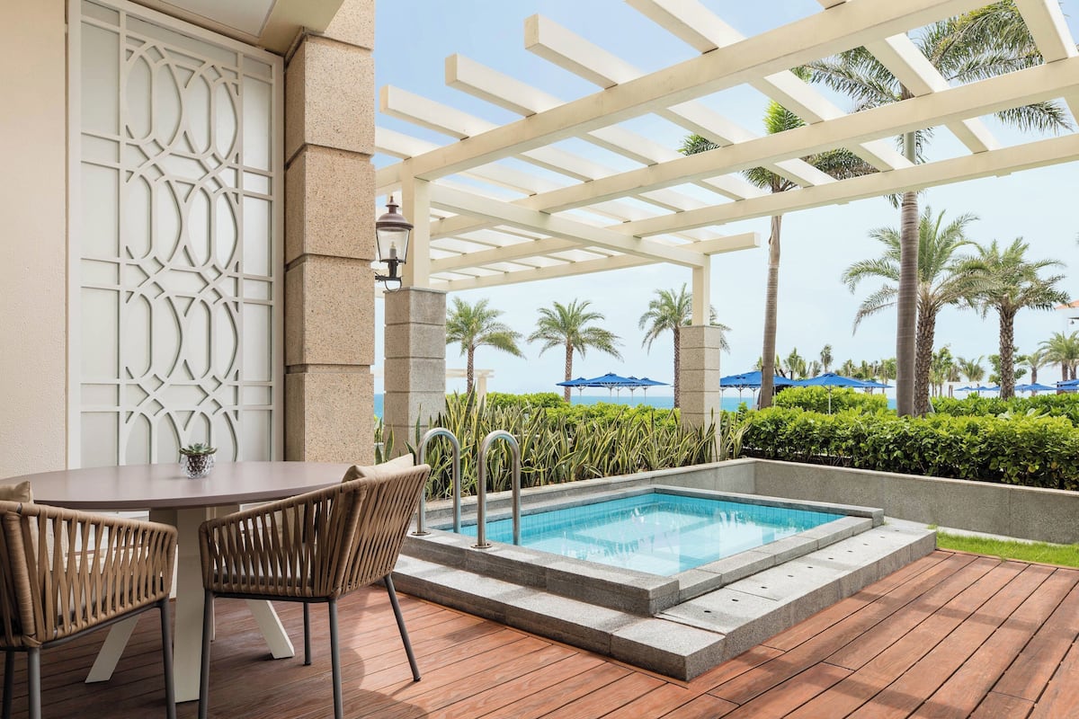 A pool area with a pergola, a table with chairs, and a view of palm trees and umbrellas.