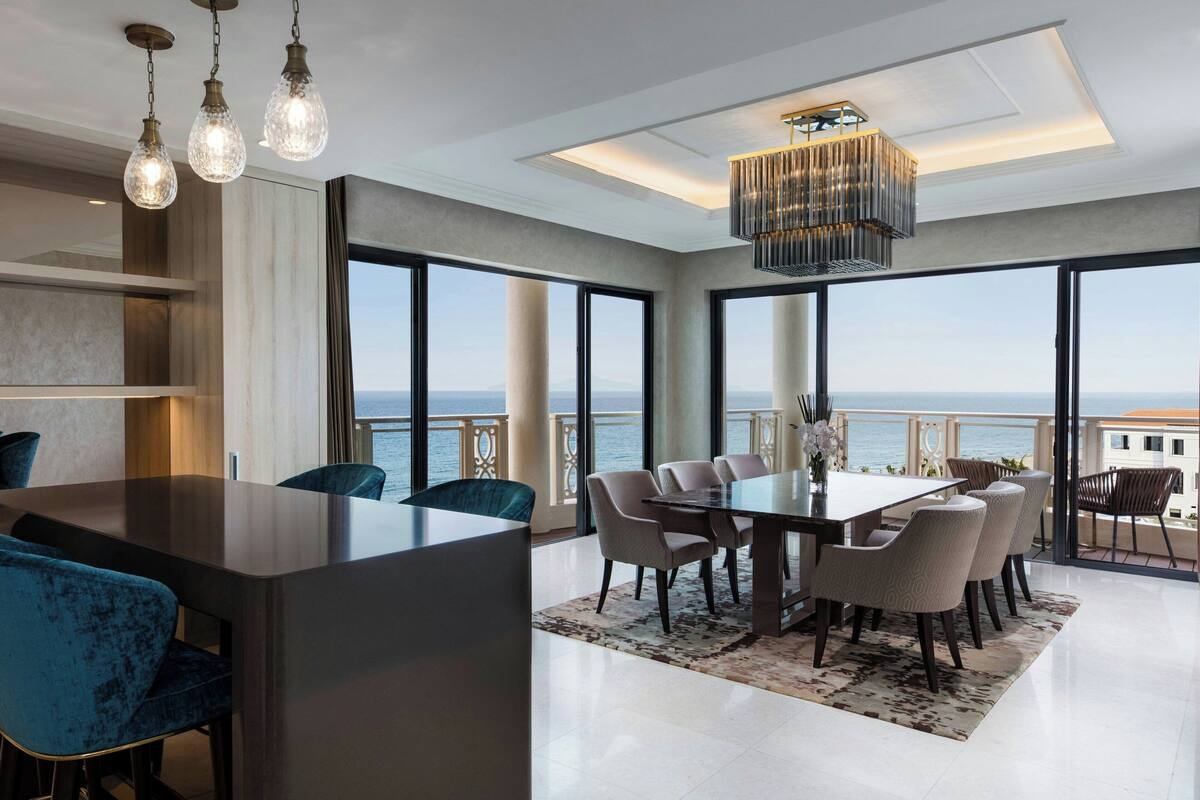A modern dining area with a large table and chairs, large windows with a view of the sea, and a chandelier.