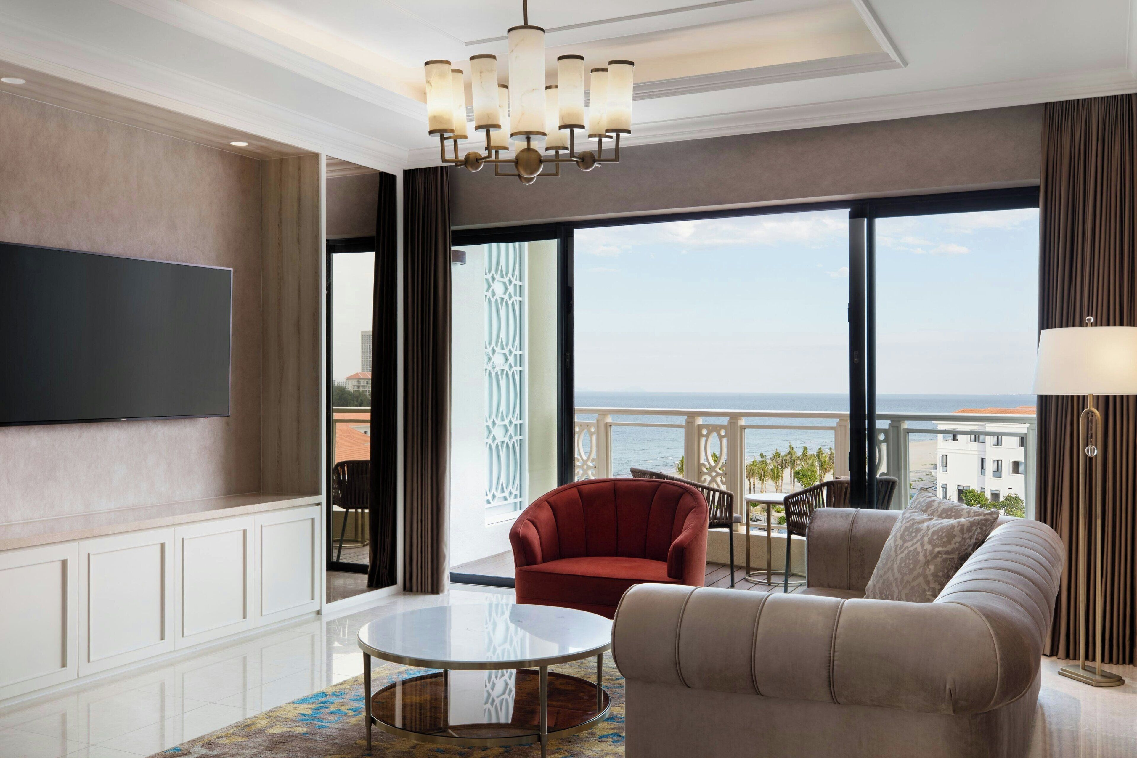 A spacious living room with a large flat-screen TV, a round coffee table, a sofa, and a red armchair. There is a balcony with a view of the ocean and buildings.
