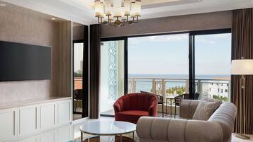 A spacious living room with a large flat-screen TV, a round coffee table, a sofa, and a red armchair. There is a balcony with a view of the ocean and buildings.