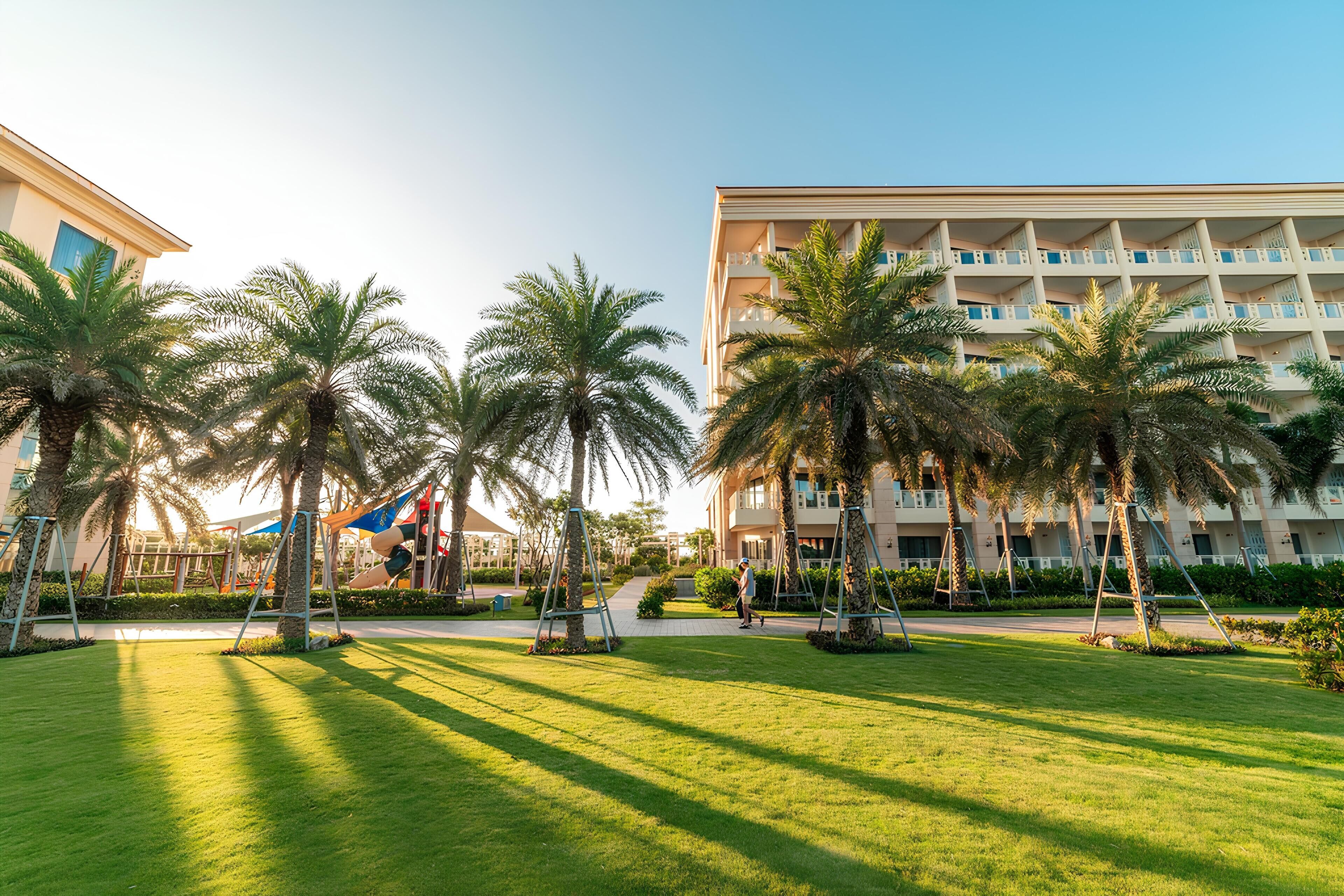 Un patio de hotel con palmeras, un parque infantil y un edificio con varios balcones.