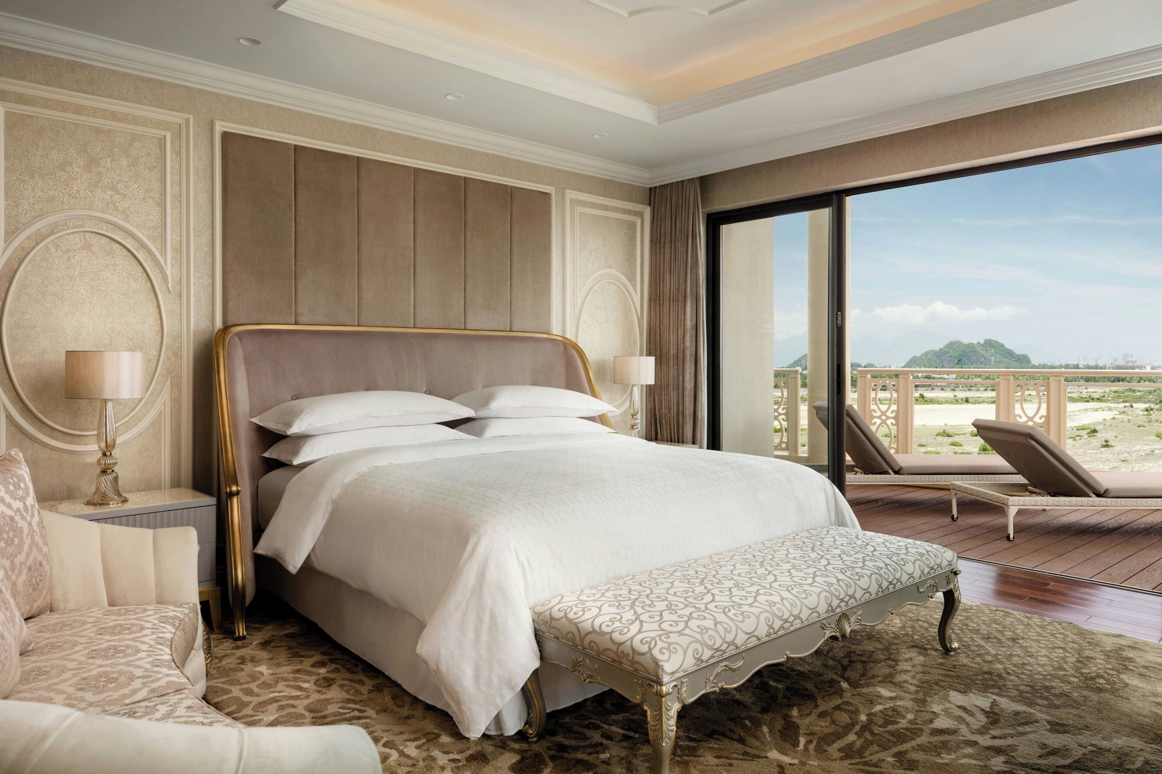 A spacious bedroom with a large bed, a bench, and a view of a balcony with lounge chairs.
