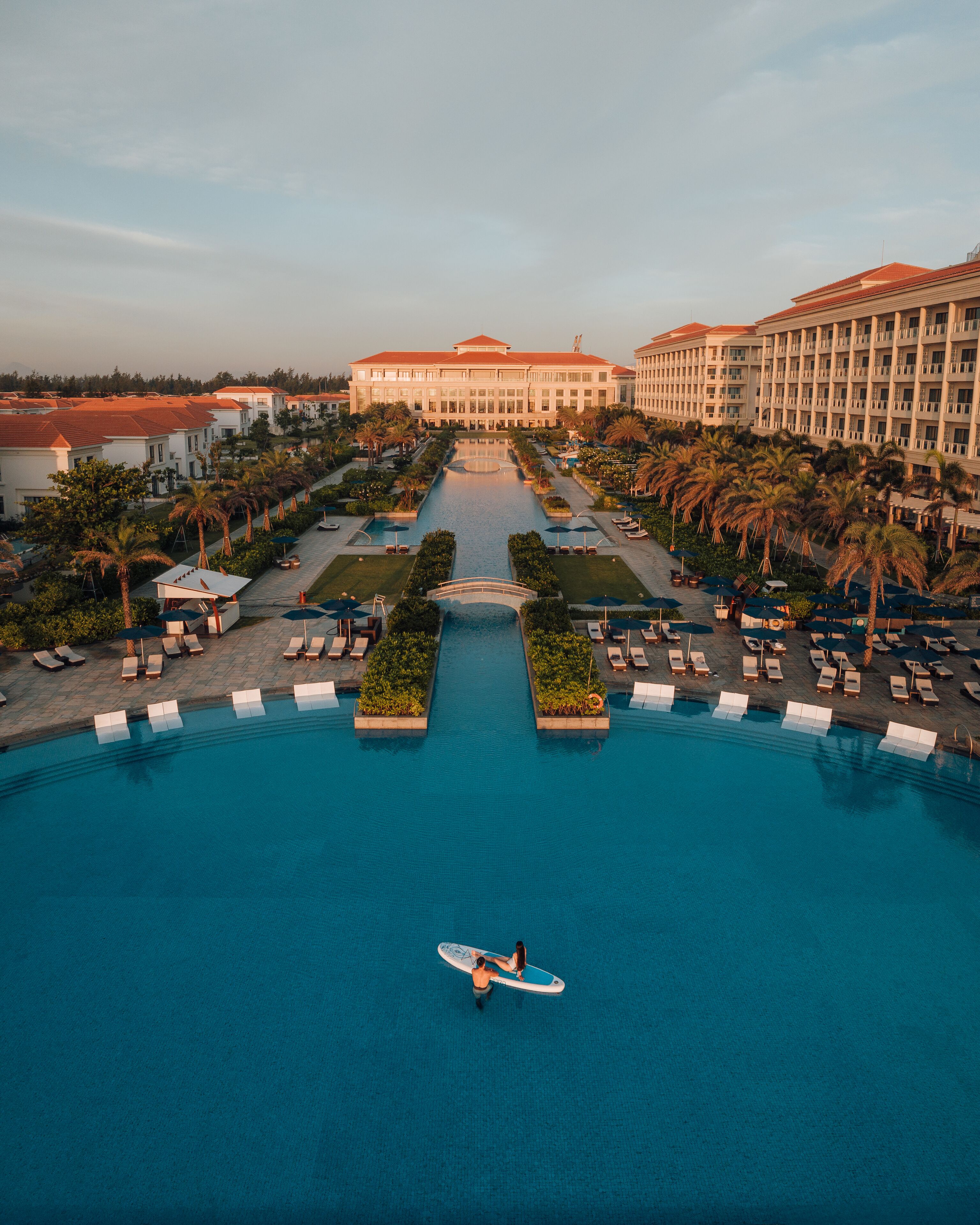 Un hotel grande con piscina central, varias edificaciones y palmeras.