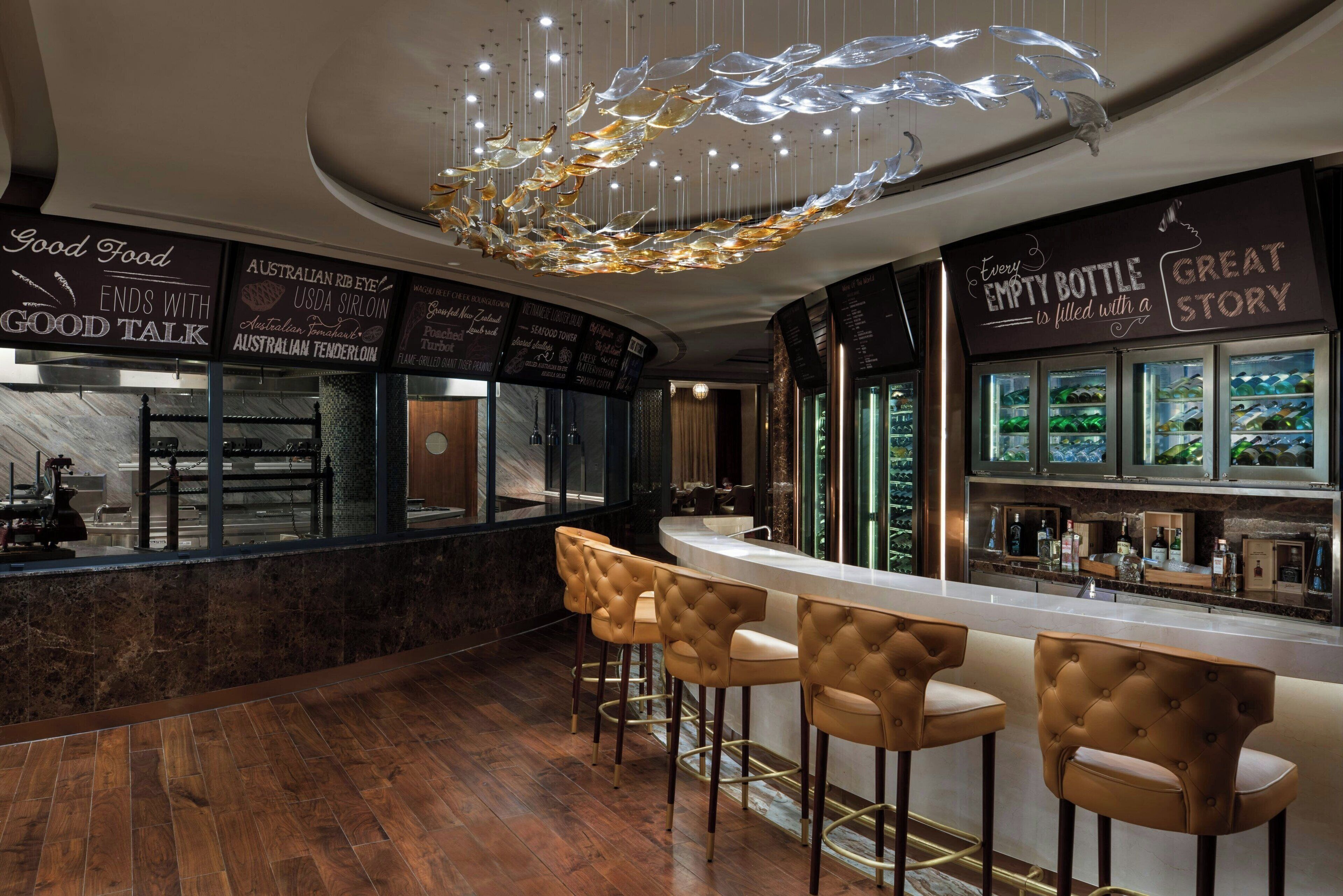A modern bar area with a marble countertop, leather bar stools, and a decorative glass chandelier.