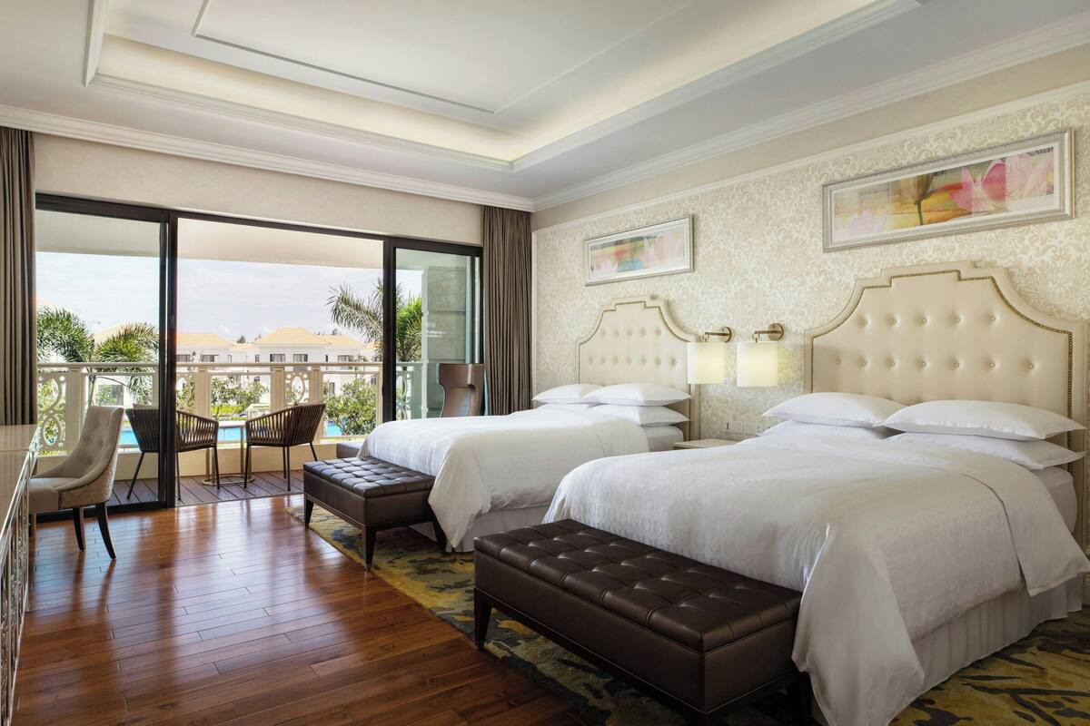 A hotel room with two beds, a sitting area with a chair and a table, and a balcony with a view of buildings and greenery.