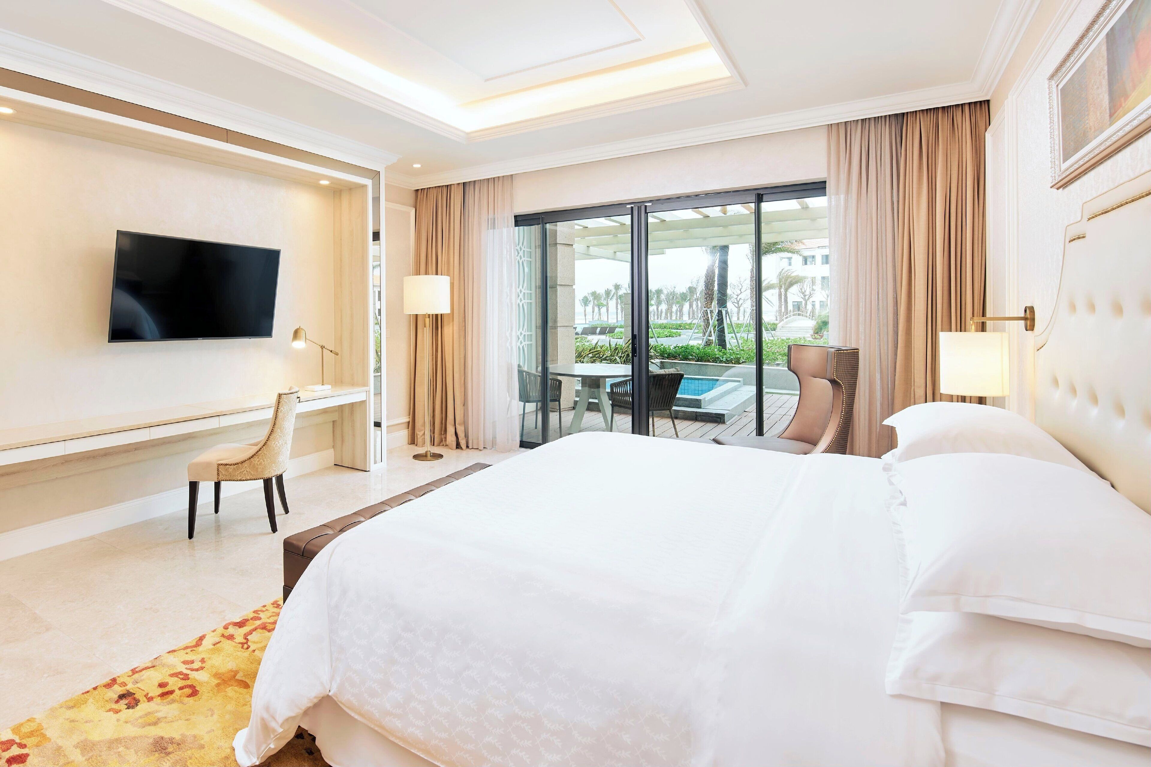 A hotel room with a large bed, a flat-screen TV mounted on the wall, a desk with a chair, and a balcony with a table and chairs.