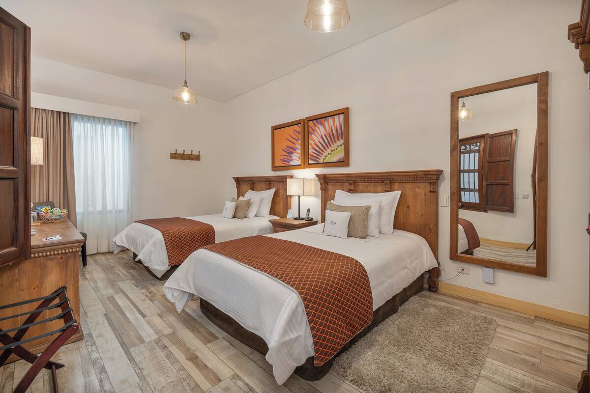 Ein Hotelzimmer mit zwei Betten, hölzernen Kopfteilen, einem Schreibtisch mit Stuhl, einem großen Spiegel und Bildern an der Wand.