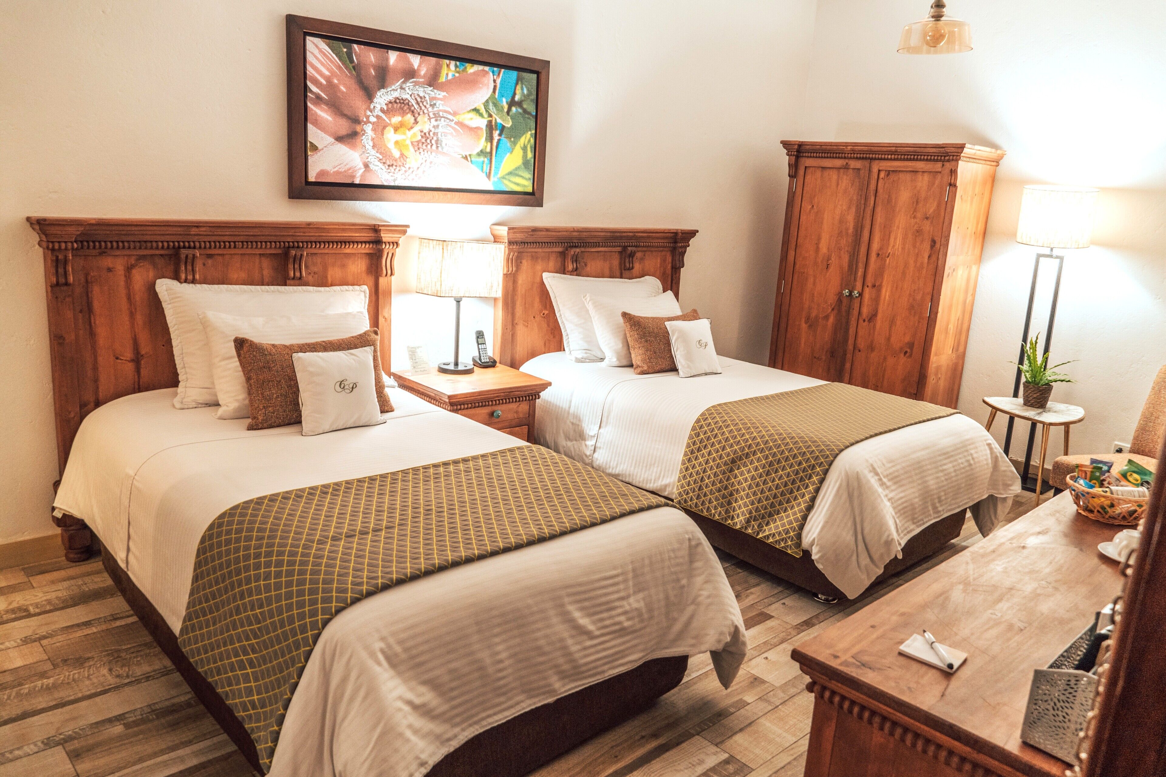 A hotel room with two beds, wooden headboards, a nightstand with a lamp, a small table with a basket, and a framed picture on the wall.