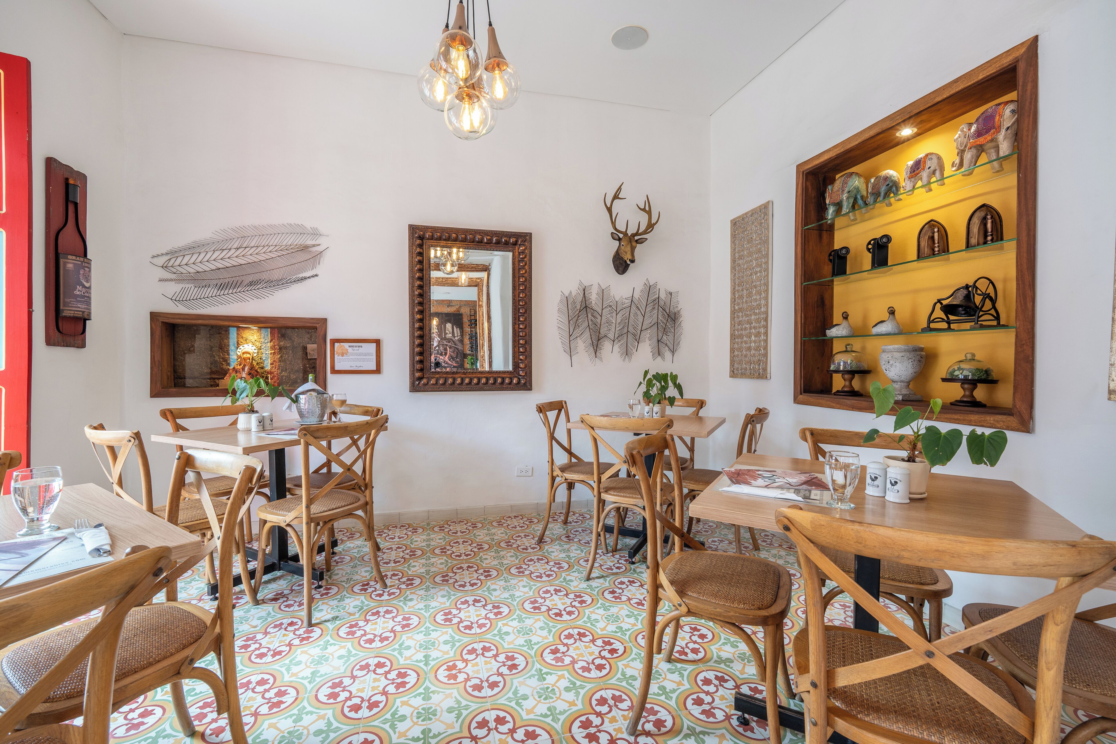 Un comedor con mesas y sillas de madera, piso estampado y decoración de pared que incluye una cabeza de ciervo y adornos con plumas.