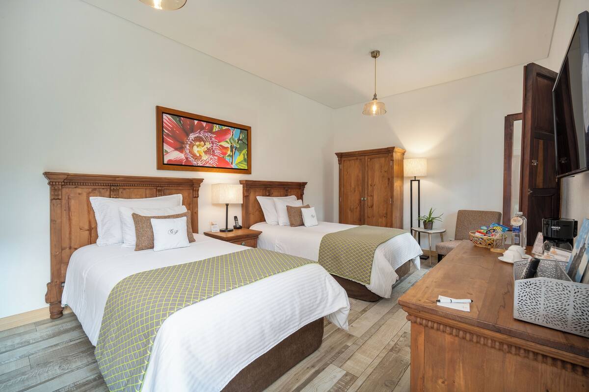 Ein Hotelzimmer mit zwei Betten, einem hölzernen Kopfteil, einem Schreibtisch mit Fernseher, einer Lampe und einem Bild an der Wand.