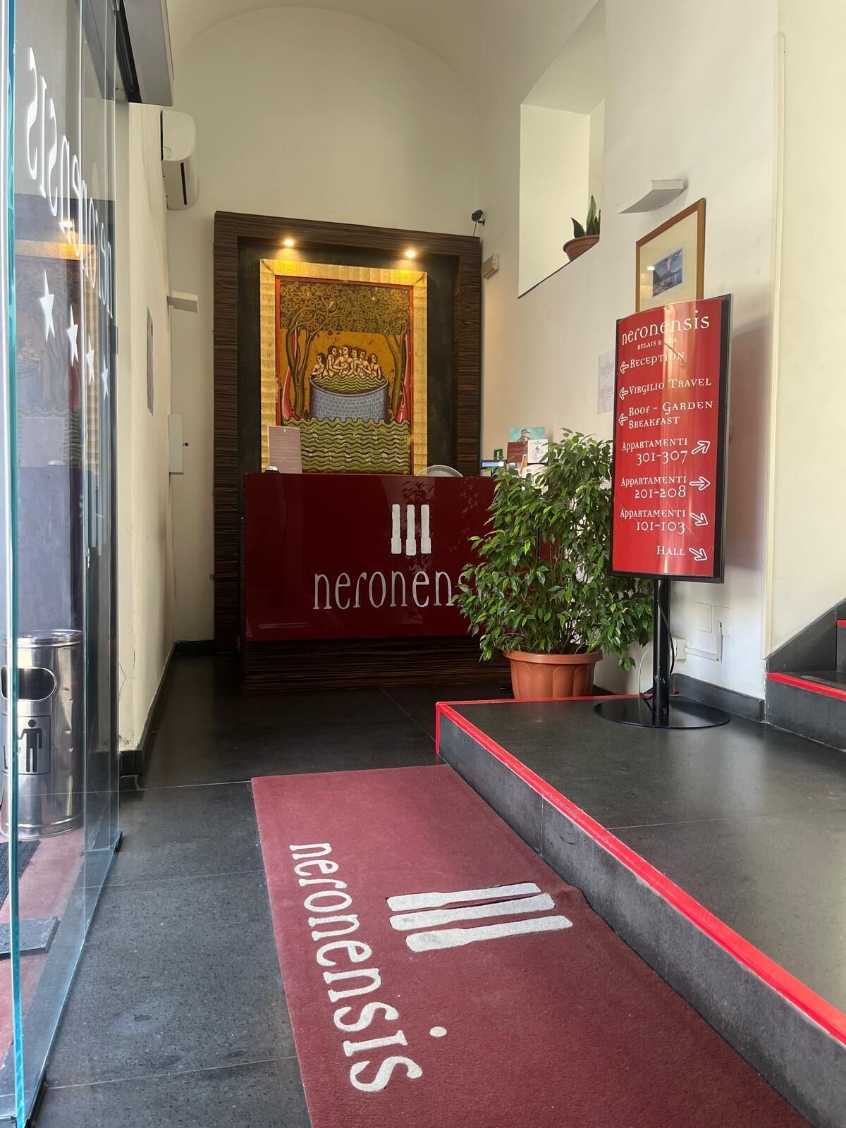 A hotel reception area with a red carpet, a reception desk, a wall-mounted sign with room numbers, and a large mural.
