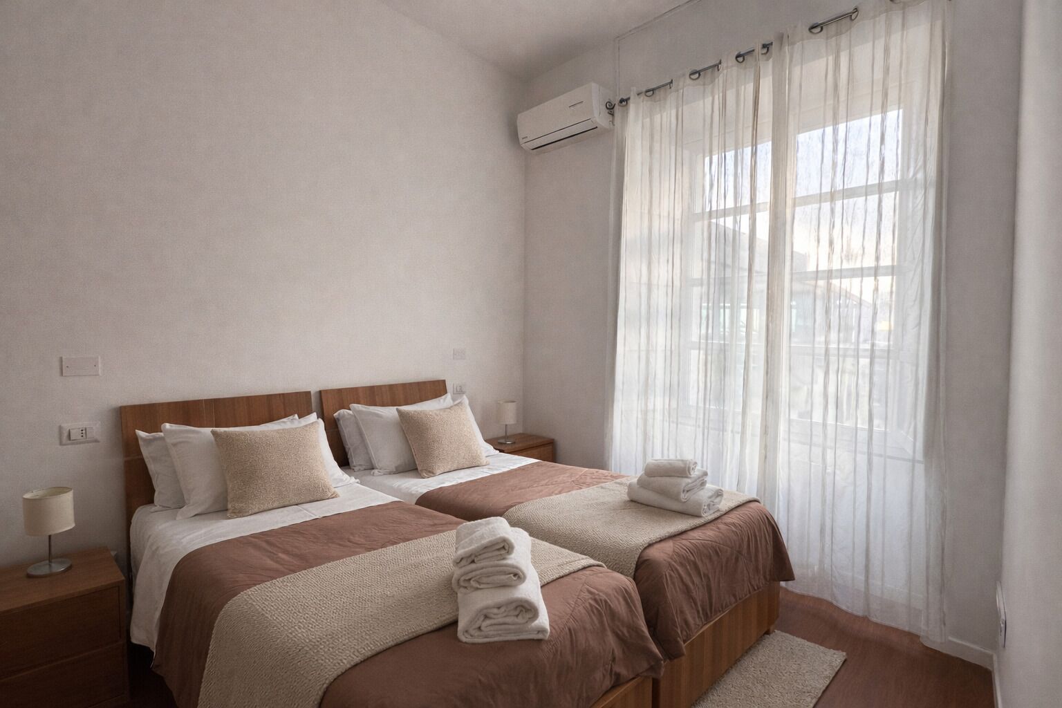 Un letto matrimoniale con testiera in legno, un comodino con lampada e una finestra con tende trasparenti.
