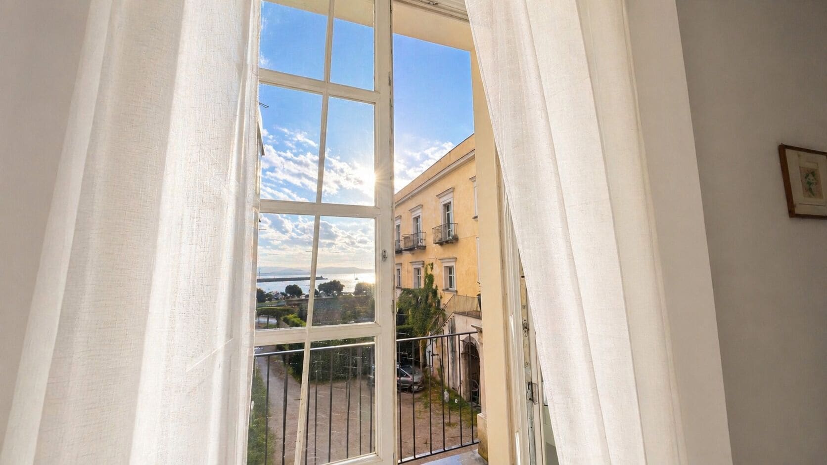 A window with white curtains, a view of a building with balconies, and a clear sky.
