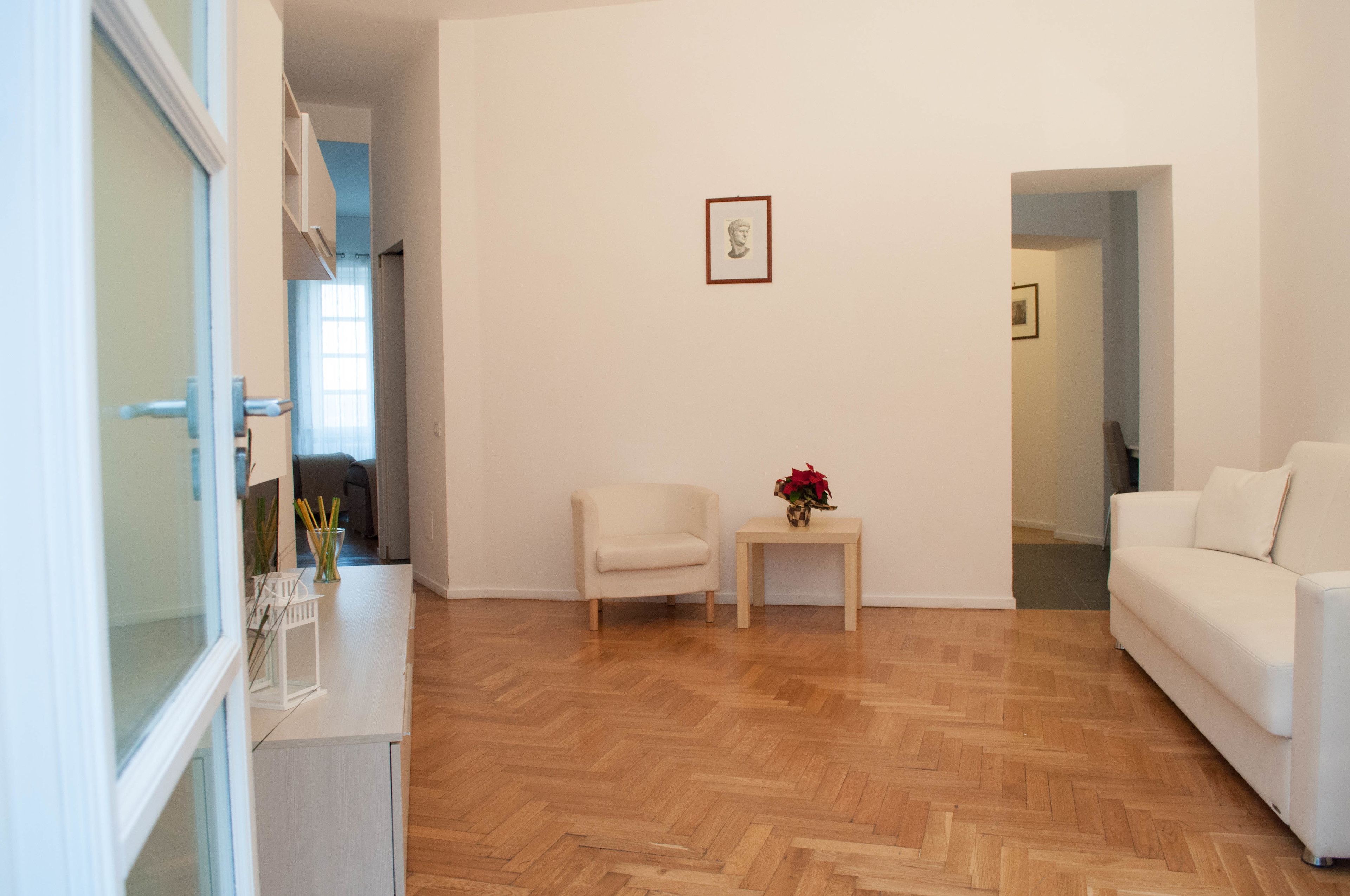 Un salon moderne avec un canapé blanc, une petite table d’appoint surmontée d’un vase et un parquet en bois à motifs chevrons.