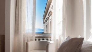 Habitació d'hotel amb un llit, una cadira i vista a un edifici i al mar a través de cortines translúcides.